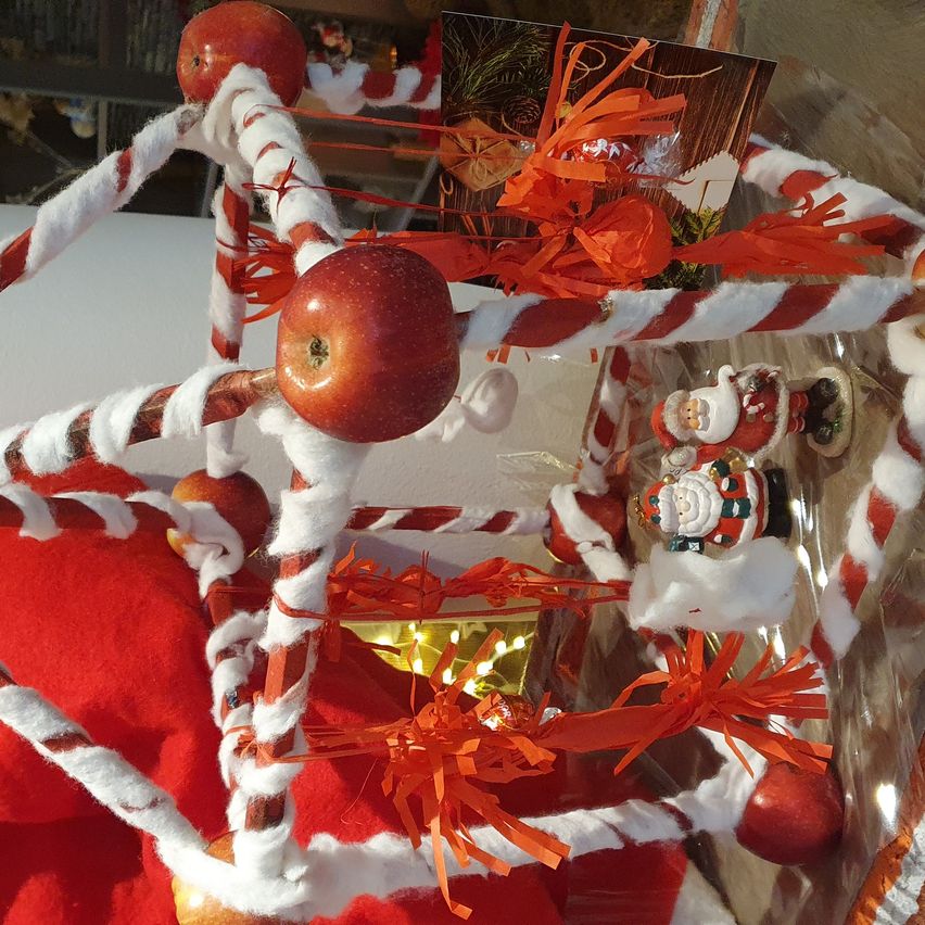 Eine Weihnachtsdekoration mit Äpfeln, weißen und roten Bändern und Weihnachtsmann-Figuren. Die Dekorationen stehen auf einer roten Basis, mit Lichtern und Ornamenten drumherum.