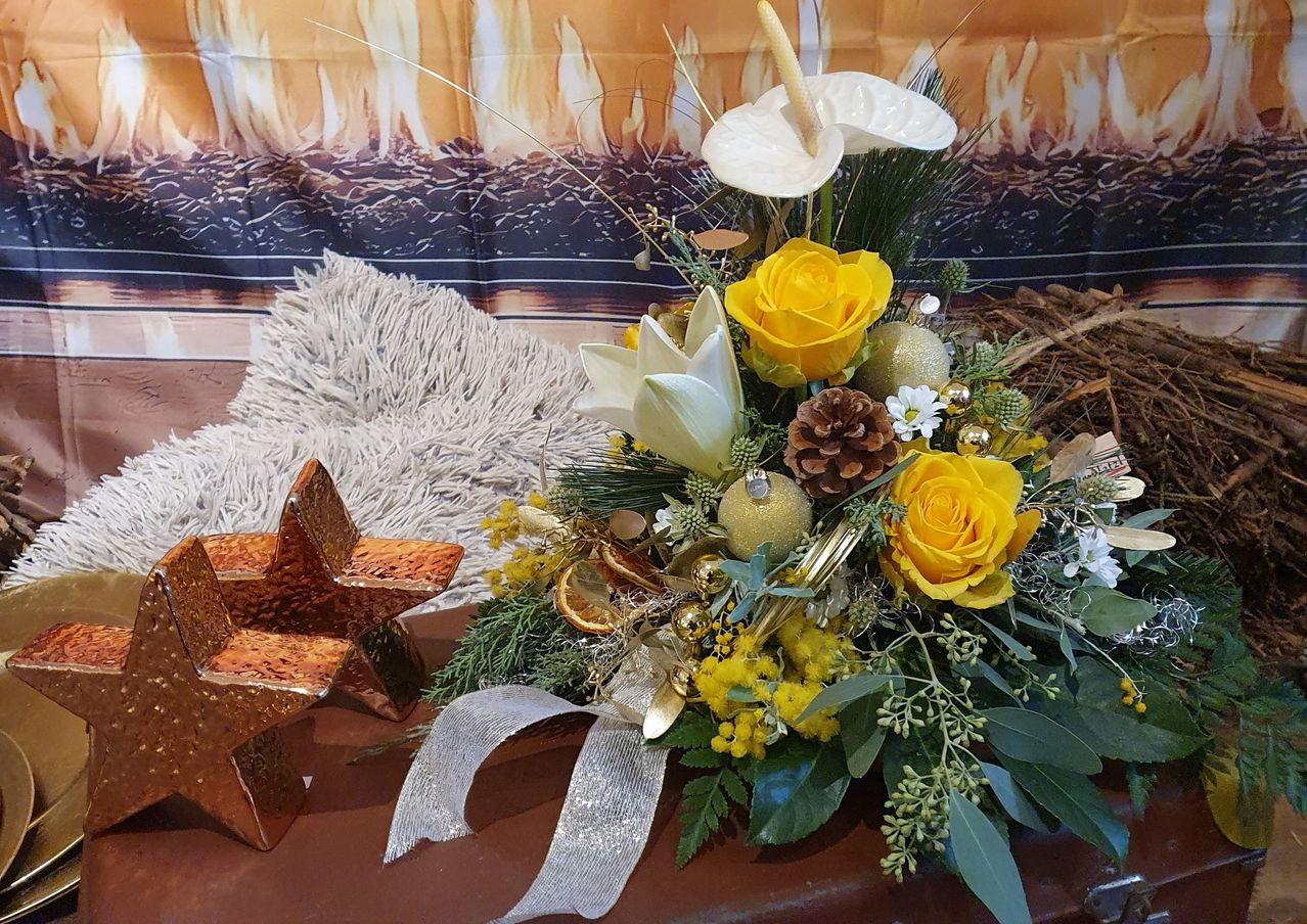 Ein weihnachtliches Blumenarrangement mit gelben Rosen, weißen Lilien und grünen Blättern. Es ist mit goldenen Ornamenten, Tannenzapfen und silbernen Bändern geschmückt. Ein Stern und ein Pelzkissen sind in der Nähe.