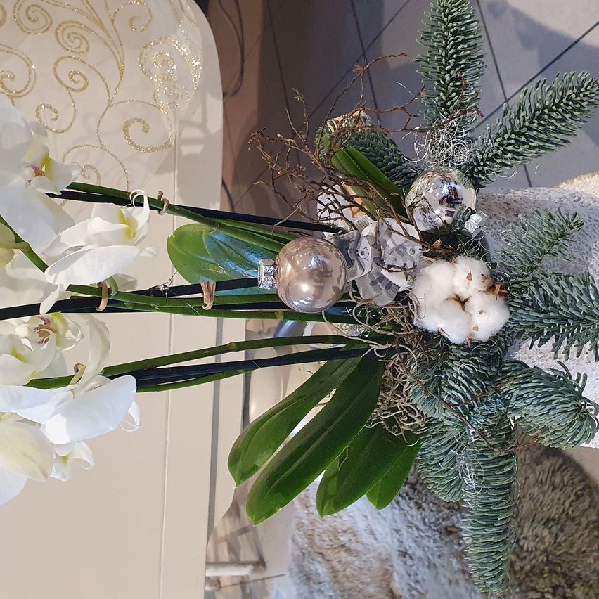 Ein Nahaufnahme einer festlichen Blumengesteckes mit weißen Orchideen, grünen Blättern, Tannenzapfen und Ornamenten. Der Hintergrund ist hell, mit einem weißen und goldenen Muster.