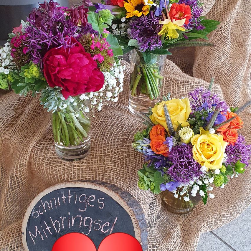 Drei Glasvasen mit farbenfrohen Blumenarrangements auf einem Jutetuch. Eine Vase hat eine rote Rose, eine andere mit lila Blumen und die letzte mit gelben und orangefarbenen Blumen. Ein Schild mit 'Schnättiges Mitbringsel' und roten Herzen.