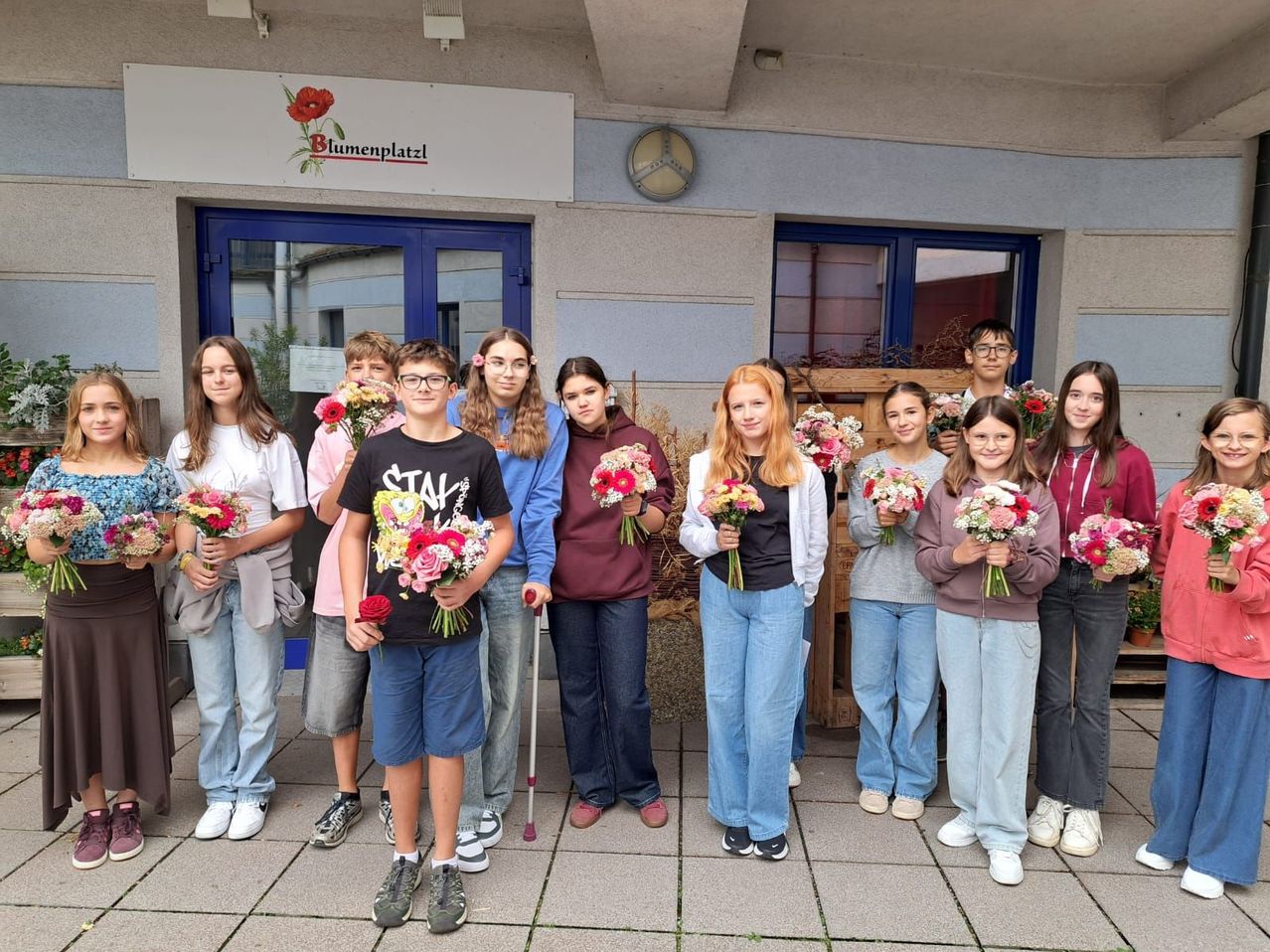 A group of young people holding flowers in front of a building with a sign that reads Blumenplatzl.