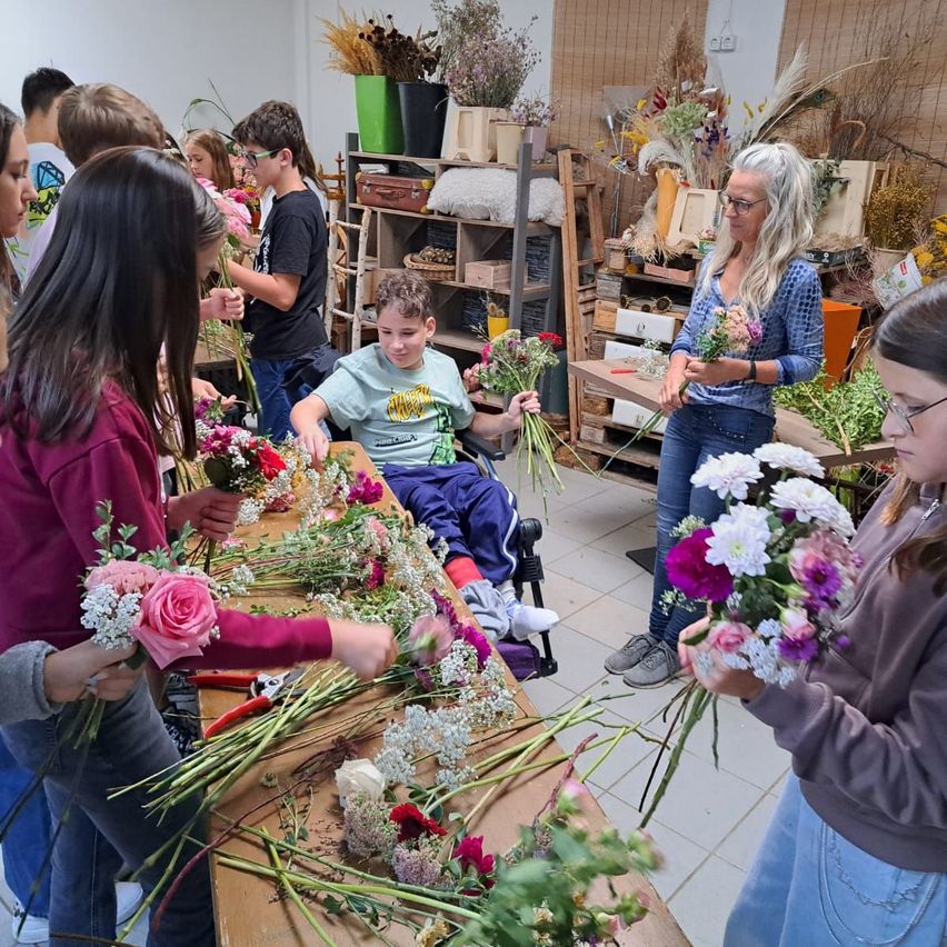 A group of people, including children, are arranging flowers in a workshop. A woman in a blue shirt is instructing them.