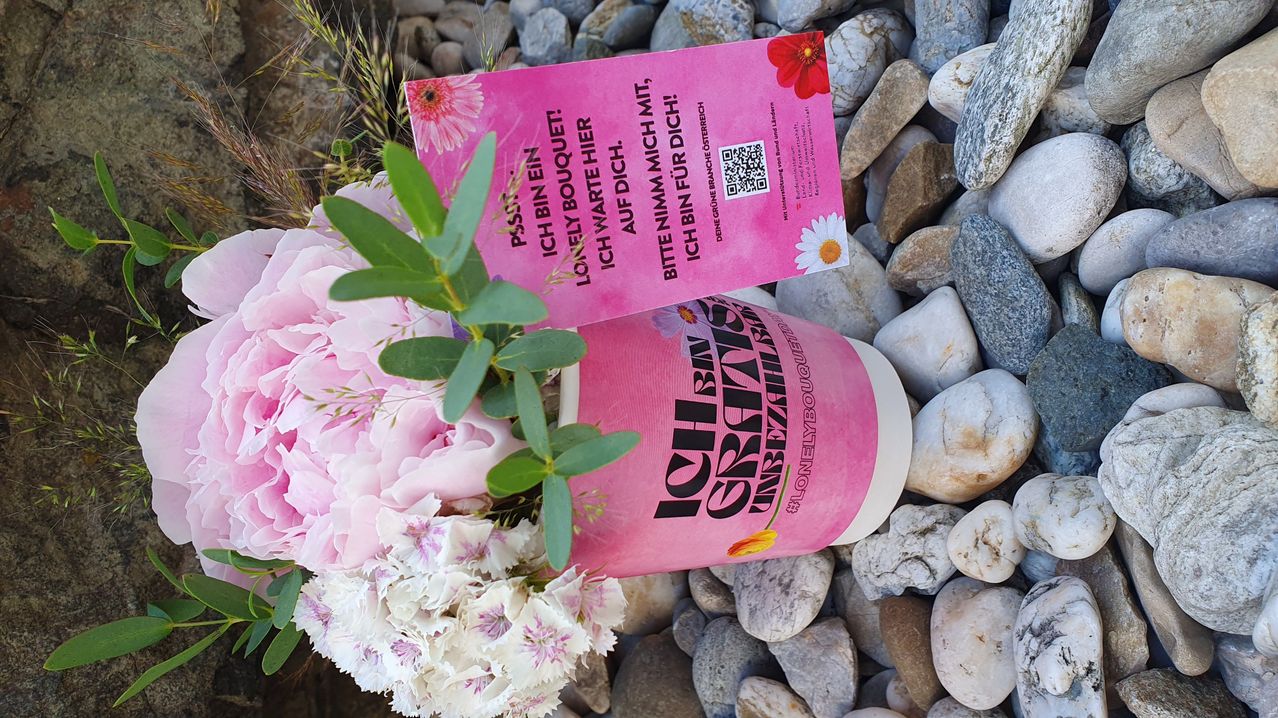 Ein rosa Blumenstrauß mit weißen Blüten und rosa Blütenblättern liegt auf Kieselsteinen. Eine rosa Karte mit schwarzem Text und einem QR-Code liegt auf dem Strauß. Auf der Karte steht 'Ich bin ein einsamer Strauß.' Darunter befindet sich auch ein rosa Behälter mit 'Ich glaub' geschrieben darauf.