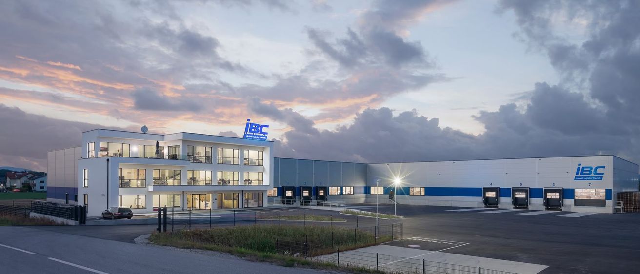 Das IBC Global Logistics Gebäude steht mit seinem Logo obenauf. Der Himmel ist teilweise bewölkt. Der Parkplatz ist eingezäunt und leer. Eine Straßenlaterne beleuchtet das Gebiet.