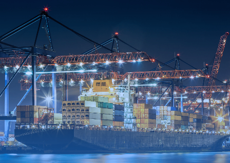 Ein Frachtschiff liegt bei Nacht unter einem Hafenkran, beleuchtet von hellen Scheinwerfern. Container sind auf dem Schiff gestapelt, und Kräne befinden sich auf beiden Seiten.