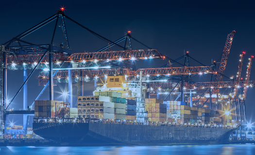 Ein Frachtschiff liegt bei Nacht unter einem Hafenkran, beleuchtet von hellen Scheinwerfern. Container sind auf dem Schiff gestapelt, und Kräne befinden sich auf beiden Seiten.