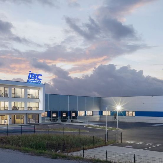 Ein großes Lagergebäude mit dem IBC-Logo oben, unter einem teilweise bewölkten Himmel. Das Gebäude hat ein modernes Design mit Glasfenstern und einem eingezäunten Parkplatz.