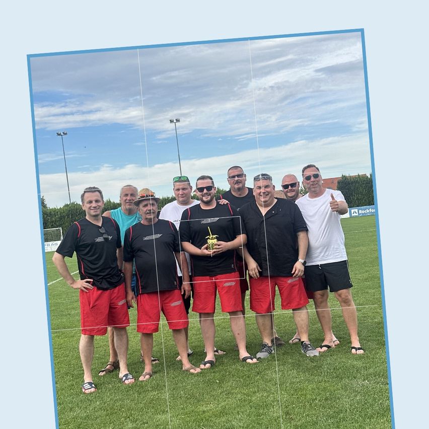 Eine Gruppe von Männern in Sonnenbrillen und roten Shorts steht auf einem Rasenfeld, einer von ihnen hält ein Getränk. Dahinter ist ein unscharfer Blick auf ein Feld mit Bäumen und einem Zaun.