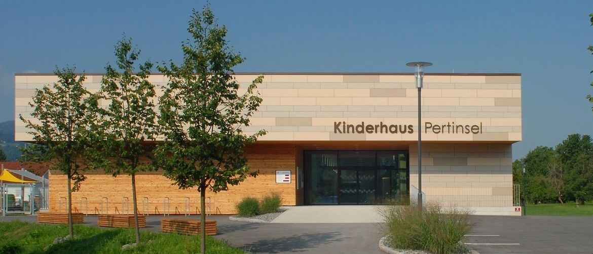 Der Eingang zum Kinderhaus Pertinsel, ein Gebäude mit Glastüren und einer Ziegelfassade, hat Bäume und eine Straßenlaterne vor sich. Der Parkplatz befindet sich rechts.