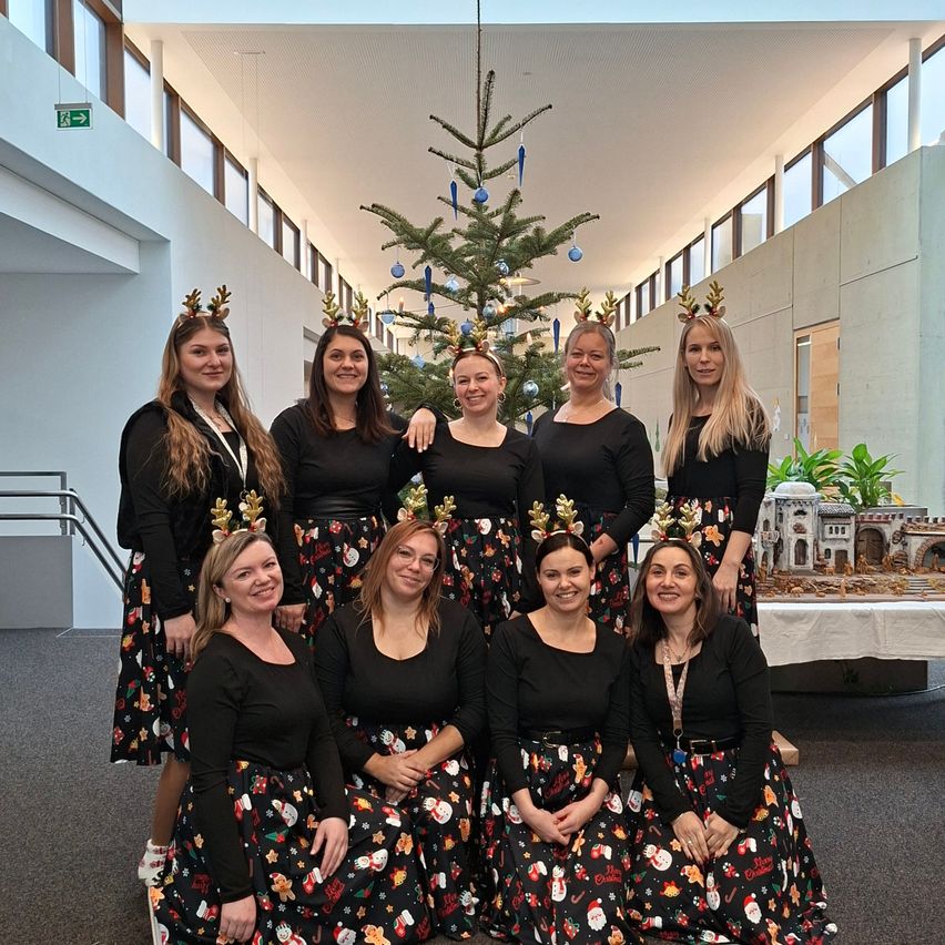 Eine Gruppe von Frauen in festlichen Rentierkopfbändern und weihnachtlich gestalteten Kleidern posiert für ein Foto vor einem Weihnachtsbaum.
