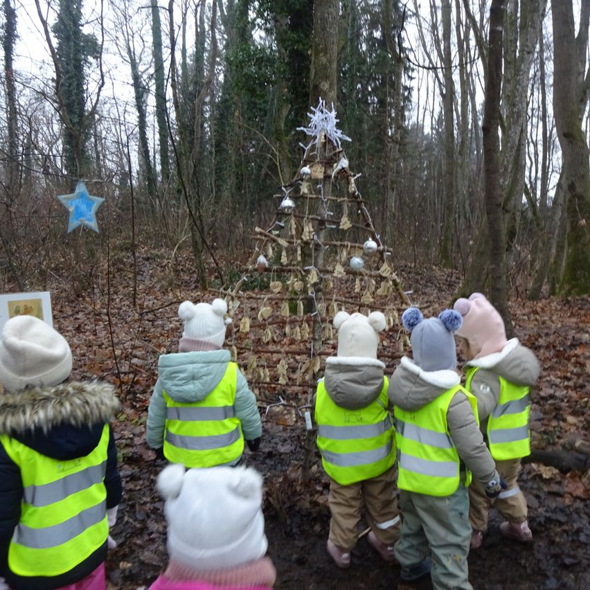 Eine Gruppe von Kindern in Warnwesten steht vor einem aus Zweigen gebauten Weihnachtsbaum im Wald. Sie tragen Winterkleidung und Mützen.