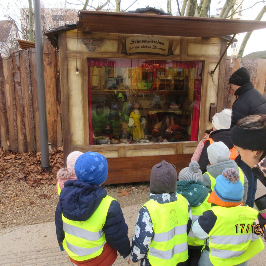 Eine Gruppe von Kindern in Sicherheitswesten betrachtet eine Weihnachtsausstellung mit sieben Zwergen in einem kleinen Holzhäuschen.