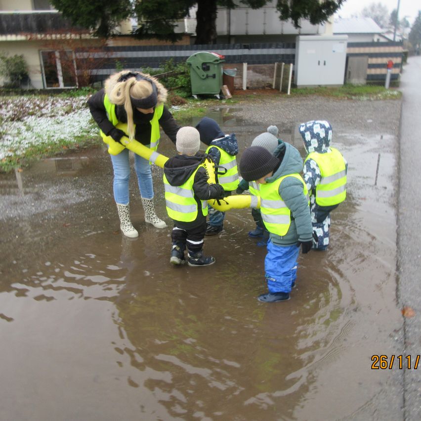 Eine Frau und mehrere Kinder in gelben Westen stehen in einer Pfütze. Sie scheinen sich gegenseitig beim Waten durch das Wasser zu helfen.