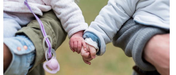 Zwei Babys halten Händchen, während sie von Erwachsenen gehalten werden, ein Nahaufnahme-Bild. Das Baby auf der linken Seite hat einen Schnuller im Mund.