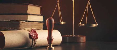 A stack of books, a scale, and a rolled-up scroll with a red wax seal are on a dark wooden table.