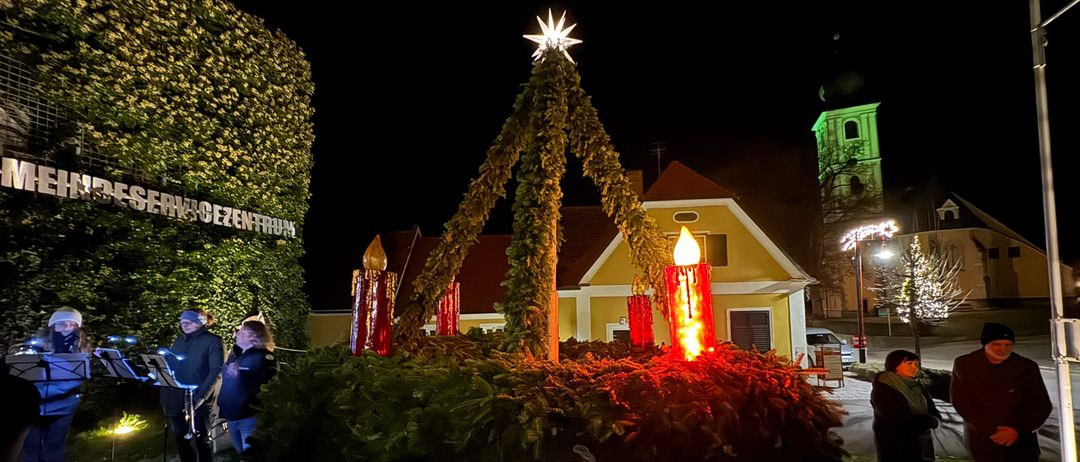 Ein großer Kranz mit roten Kerzen und einem Stern oben drauf steht vor einem Gebäude. Zwei Personen spielen vor ihm Instrumente. Ein Turm steht hinter dem Gebäude.