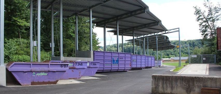 Mehrere lila Müllcontainer sind unter einem Metalldach aufgereiht, mit einer gepflasterten Straße davor. Bäume und ein Gebäude sind im Hintergrund zu sehen.