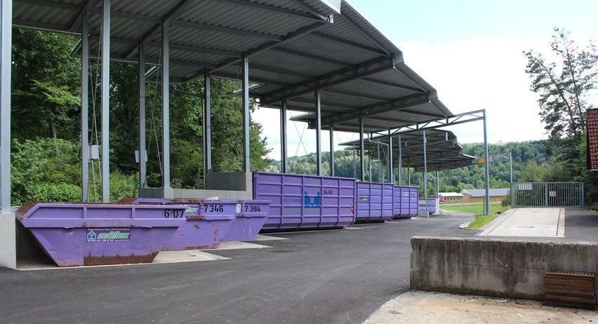 Mehrere lilafarbene Müllcontainer sind unter Metalldächern aufgereiht. Die Container haben weißen Text und Zahlen darauf. Im Hintergrund befinden sich Bäume und Büsche. Auf der rechten Seite steht ein Metallzaun.