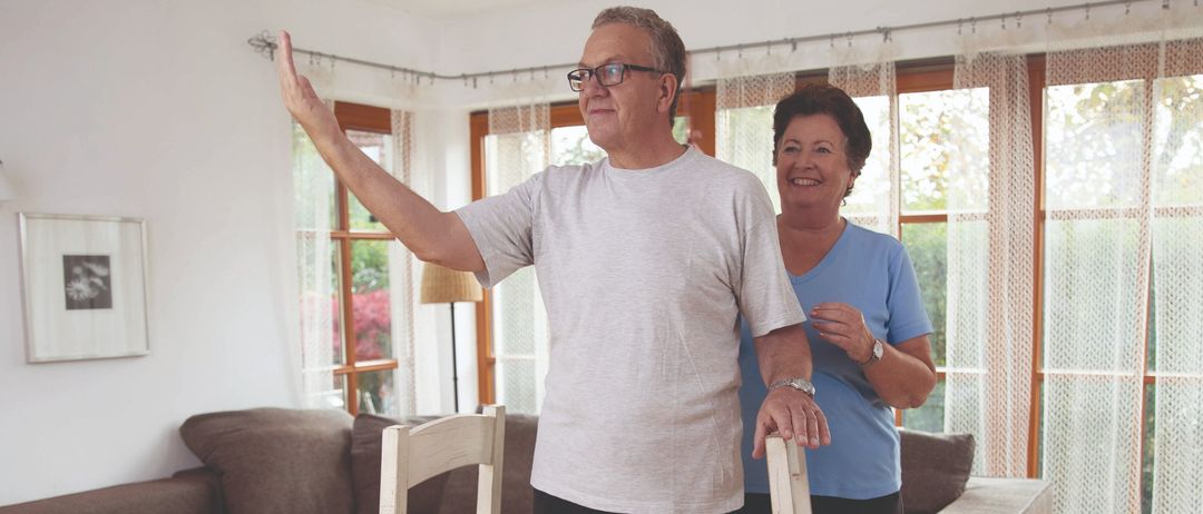 Ein älteres Paar übt das Aufstehen von einem Stuhl in einem Wohnzimmer mit Holzboden und einer braunen Couch.