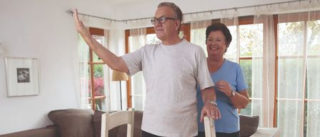 Ein älteres Paar übt das Aufstehen von einem Stuhl in einem Wohnzimmer mit Holzboden und einer braunen Couch.