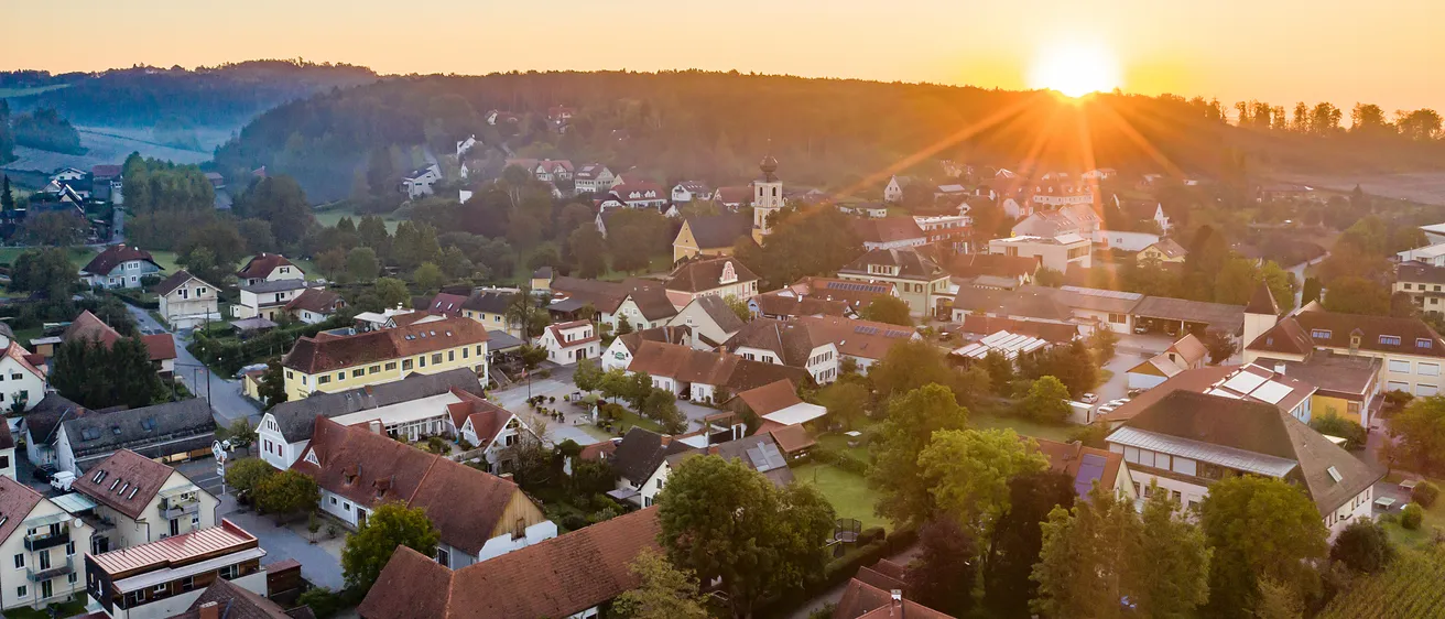 Ein Panoramablick auf ein Dorf bei Sonnenuntergang mit klarem Himmel, Häusern mit roten Dächern und einem Turm in der Ferne.