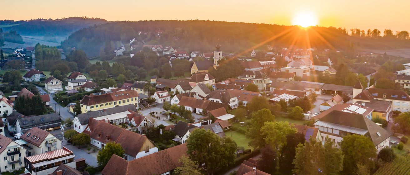 Ein Panoramablick auf ein Dorf bei Sonnenuntergang mit klarem Himmel, Häusern mit roten Dächern und einem Turm in der Ferne.