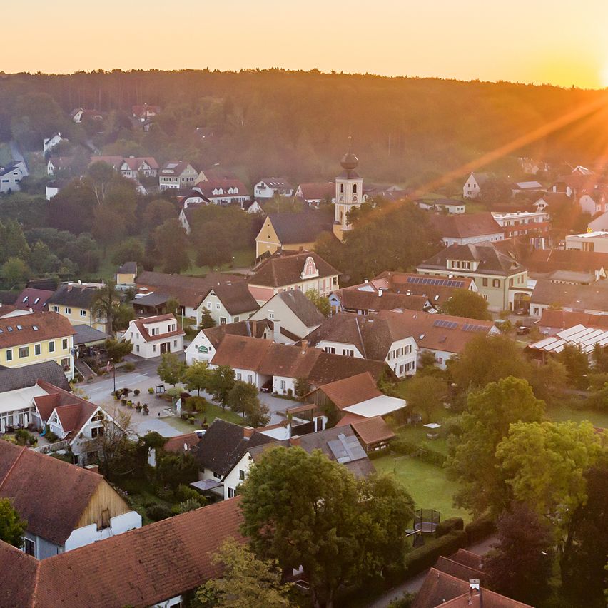 A panoramic view of a village at sunset with a clear sky, houses with red roofs, and a tower in the distance.