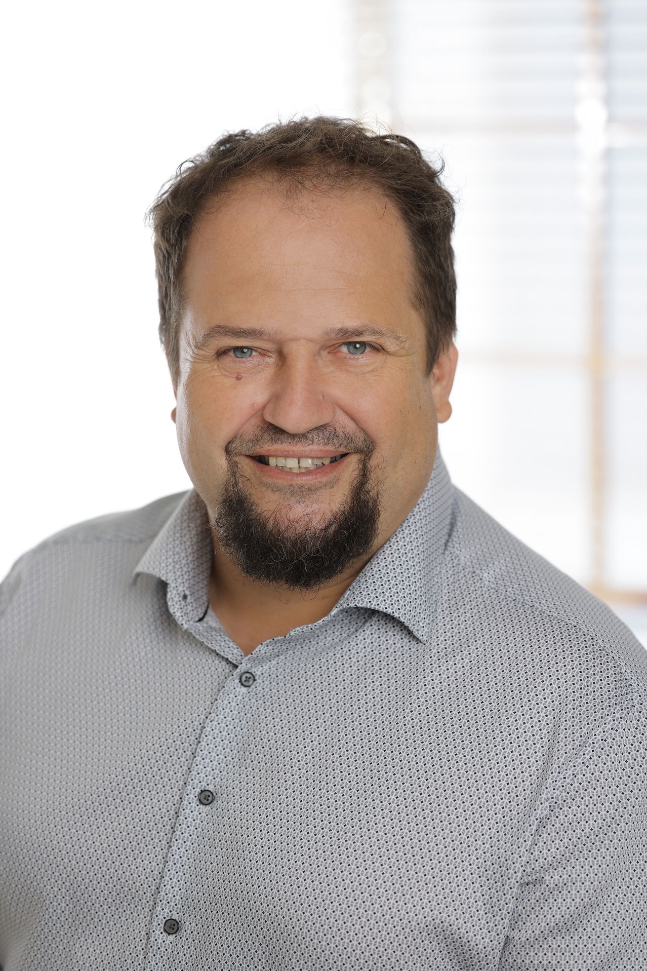 Ein Mann mit Bart lächelt und schaut in die Kamera. Er trägt ein hellgraues Hemd mit kleinen Punkten. Er befindet sich wahrscheinlich in einem Studio mit weißem Hintergrund.
