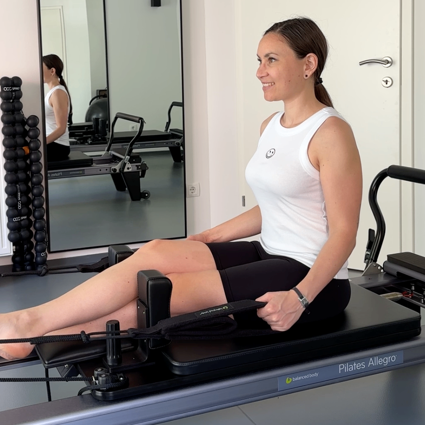 Eine Frau in einem weißen Tanktop und schwarzen Leggings trainiert auf einem Pilates-Gerät. Sie lächelt, schaut nach vorne und benutzt eine Widerstandsband. Geräte und ein Spiegel befinden sich hinter ihr.