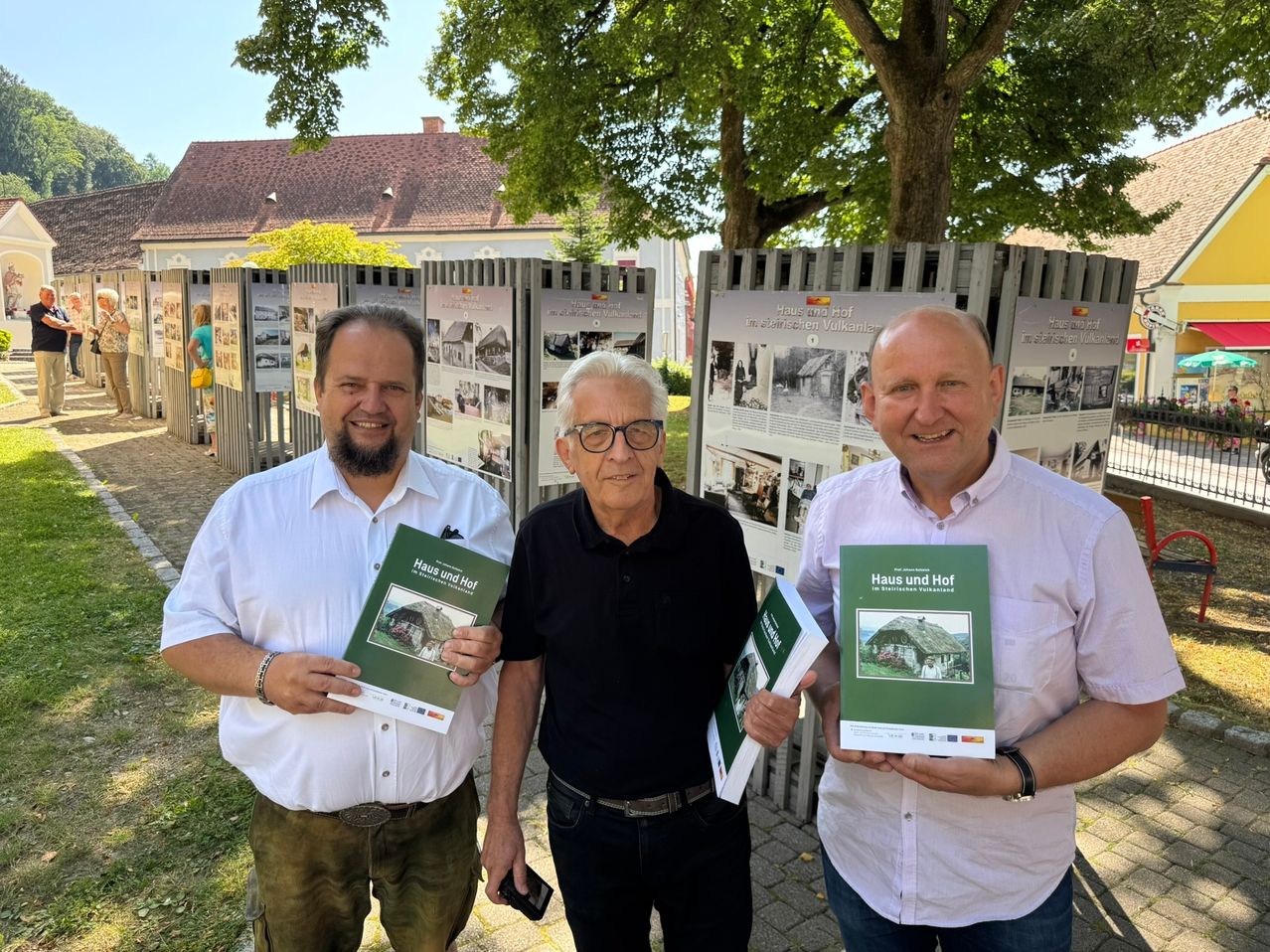 Drei Männer stehen nebeneinander und halten Bücher in den Händen. Sie sind im Freien mit Bäumen und einem Gebäude im Hintergrund. Sie alle lächeln und scheinen für ein Foto zu posieren.