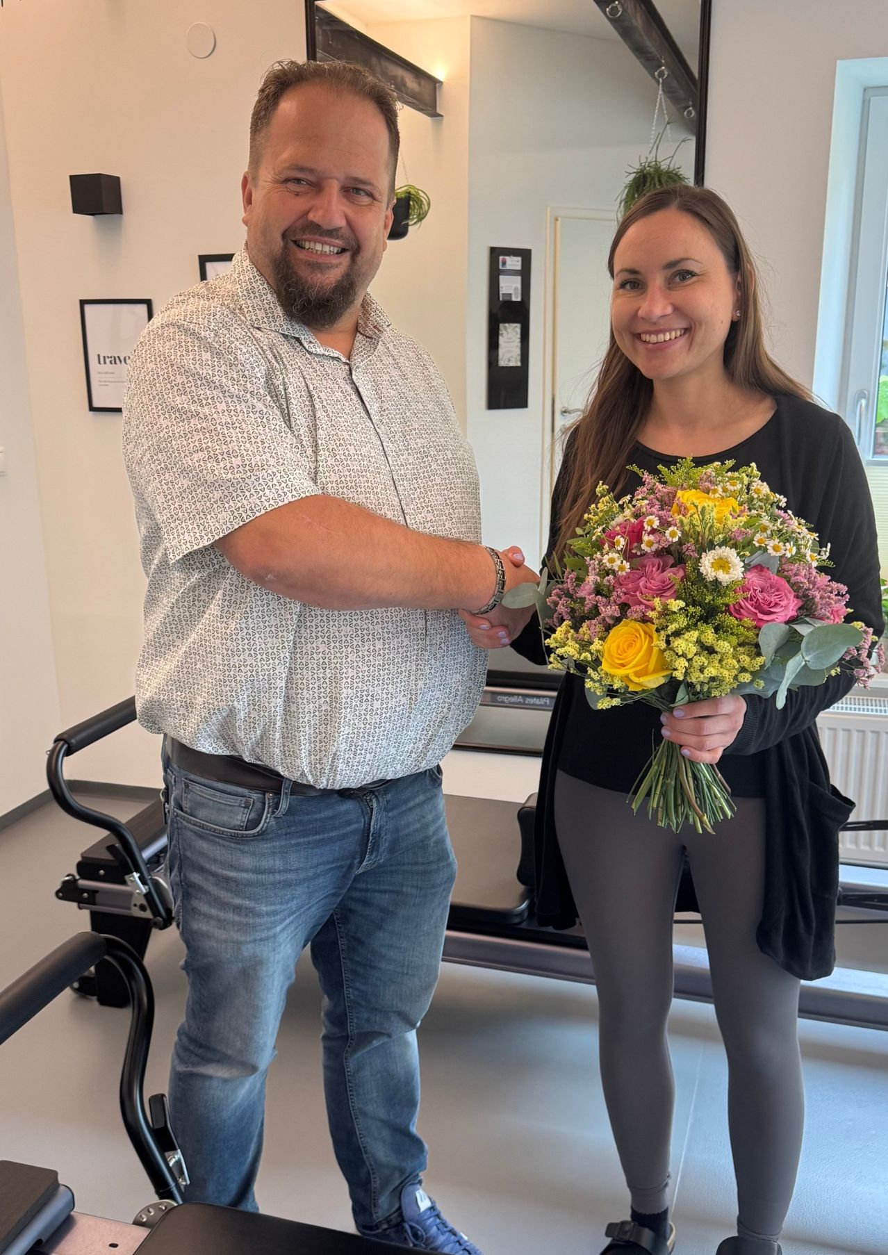 Ein Mann und eine Frau stehen lächelnd zusammen in einem Raum. Der Mann schüttelt die Hand der Frau, die einen bunten Blumenstrauß hält. Sie sind von Fitnessgeräten umgeben.