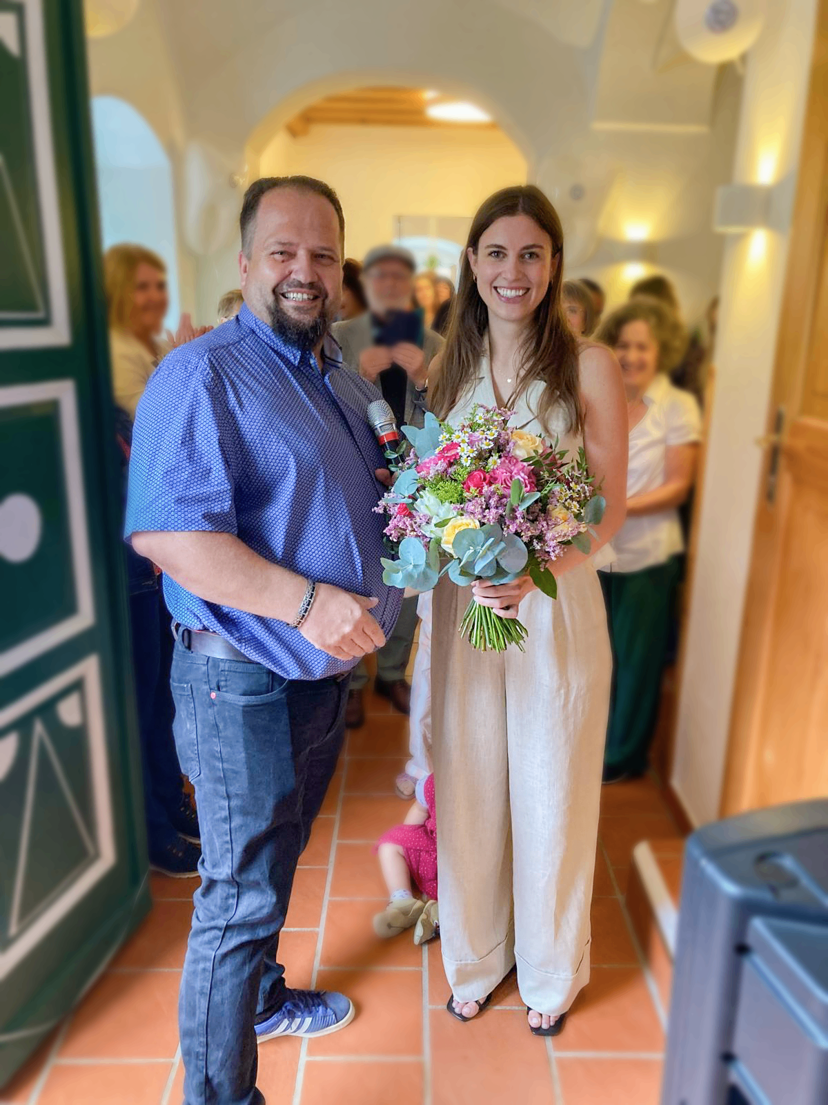 Ein Mann und eine Frau stehen zusammen, der Mann hält ein Mikrofon und einen Blumenstrauß und lächelt, während sie in die Kamera schauen. Die Frau hält den Blumenstrauß und scheint für ein Foto zu posieren. Hinter ihnen stehen mehrere Menschen und beobachten, einige halten ein Baby.