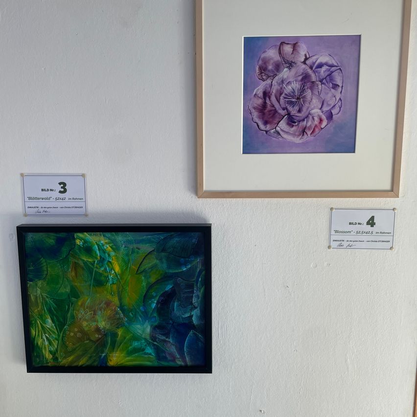Zwei gerahmte Gemälde hängen an einer weißen Wand. Das obere Gemälde ist lila mit einem Blumendesign, beschriftet mit 'Blütenwald'. Darunter befindet sich ein grünes abstraktes Gemälde, beschriftet mit 'Blossom'.