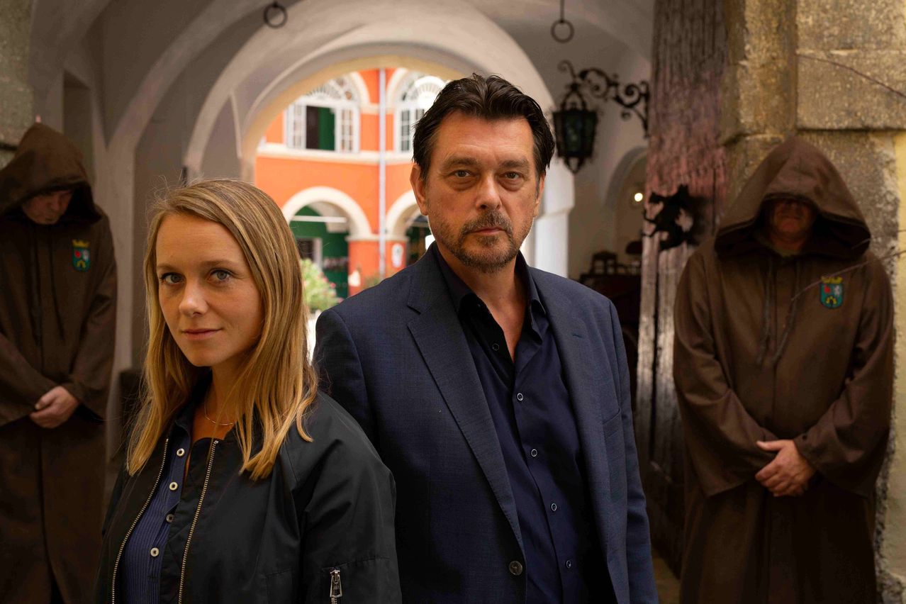 Ein Mann und eine Frau stehen gemeinsam vor der Kamera, ein orangefarbenes Gebäude und ein Bogengang im Hintergrund.