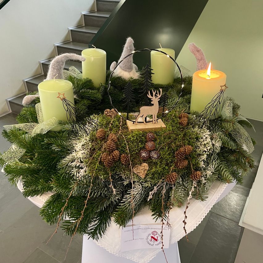 Ein Kranz aus immergrünen Zweigen mit vier grünen Kerzen, einem hölzernen Hirsch und Tannenzapfen. Er steht auf einem weißen Tisch mit einer Visitenkarte, die NZ lautet.