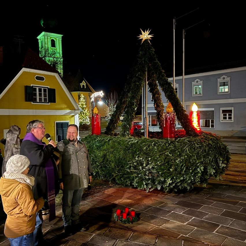 Zwei Männer stehen nachts vor einem Weihnachtsbaum, einer hält ein Mikrofon, der andere mit Bart und Brille. Ein Kind in Mütze und Schal ist in der Nähe. Ein gelbes Haus und ein Kirchturm sind im Hintergrund.
