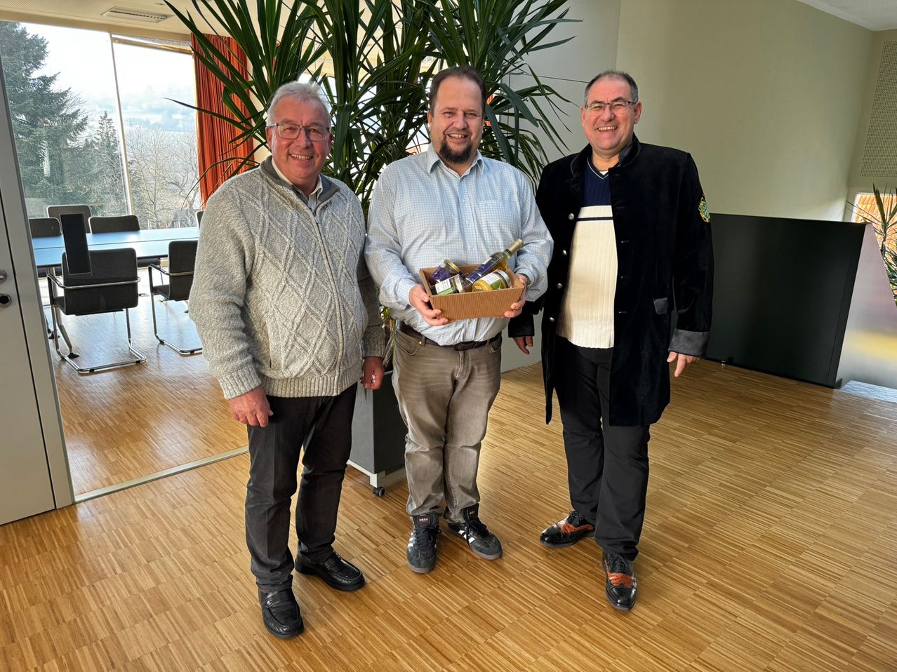 Drei Männer stehen in einem Büroraum. Der Mann in der Mitte hält eine Box mit Weinflaschen. Sie alle lächeln.