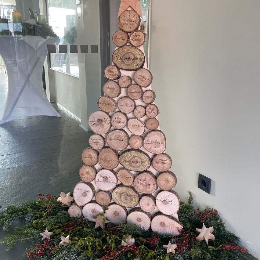Ein aus Baumstämmen gefertigter Weihnachtsbaum mit Namen steht in einem Flur mit Glaswand, umgeben von Grün und roten Beeren.