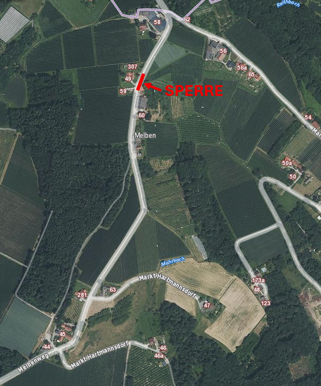 Eine Luftaufnahme einer ländlichen Landschaft mit verschiedenen Grundstücken, die mit Zahlen beschriftet sind. Der Hauptfokus liegt auf einem Bereich, der mit 'SPERRE' beschriftet ist, auf den ein roter Pfeil zeigt. Andere Beschriftungen umfassen 'Melben', 'Markt Hartmannsdorf' und mehrere nummerierte Grundstücke. Bäume und Büsche sind über die Landschaft verstreut.