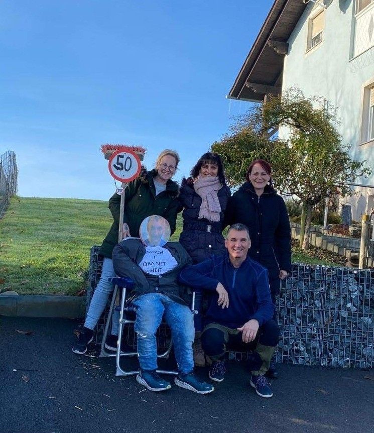 Fünf Personen posieren für ein Foto neben einem 50 km/h Geschwindigkeitsschild. Eine Person sitzt in einem Stuhl und trägt einen Papierkopf mit der Aufschrift 'OBA NET HEIT'. Die Gruppe lächelt. Hinter ihnen befindet sich eine Rasenfläche und ein Gebäude.