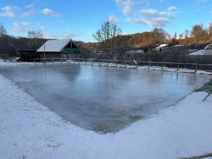 Eine gefrorene Eisbahn umgeben von Schnee und einem Zaun, mit einem Haus im Hintergrund unter einem bewölkten Himmel.