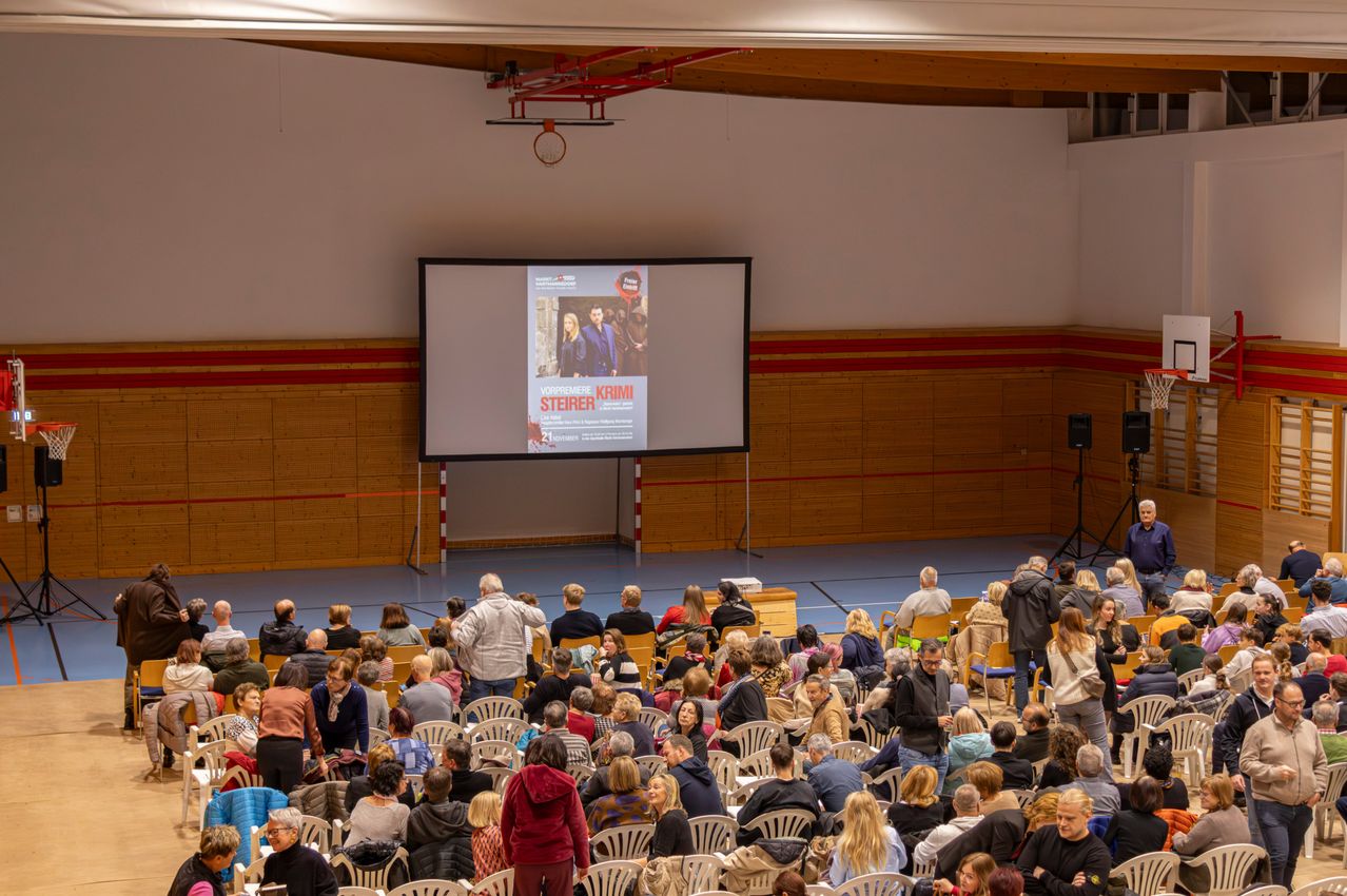 Ein großes Publikum ist in einer Halle versammelt und sieht sich einen auf einer Leinwand projizierten Film an. Das gezeigte Filmplakat ist für 'Steirerkrimi'. Die Halle hat Holzwände und einen blauen Boden.