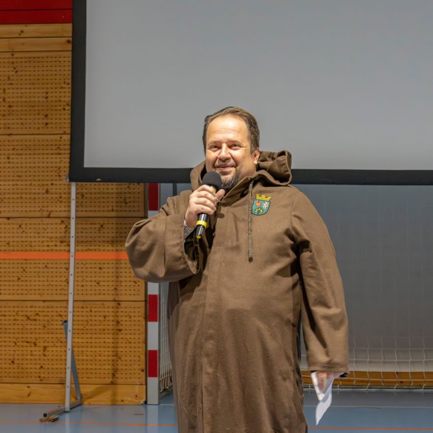 Ein Mann in einem braunen Gewand mit Kapuze steht in einer Turnhalle, hält ein Mikrofon in der Hand und lächelt, mit einer Projektionsleinwand und einer Holzwand im Hintergrund.