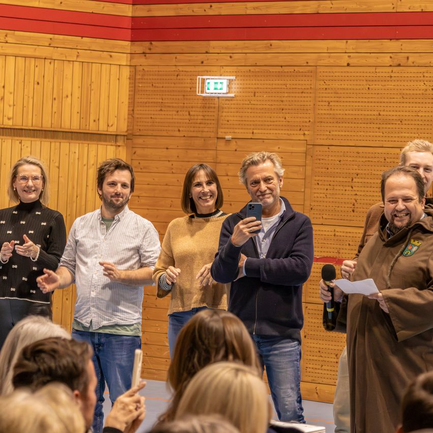 Eine Gruppe Erwachsener steht vor einer Holzwand mit einem Mikrofon in der Hand, lächelt und posiert vielleicht für ein Foto.