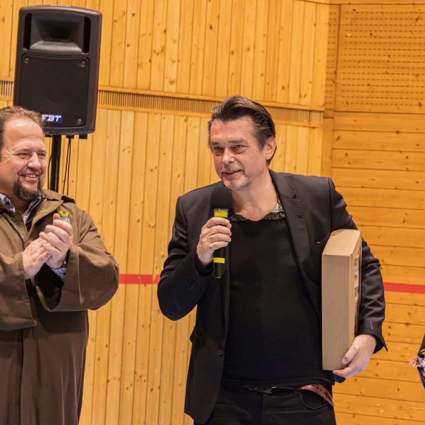 Zwei Männer stehen eng beieinander, einer hält ein Mikrofon und der andere eine Box, beide lächeln. Hinter ihnen ist eine Holzwand mit einem Lautsprecher auf einem Ständer.