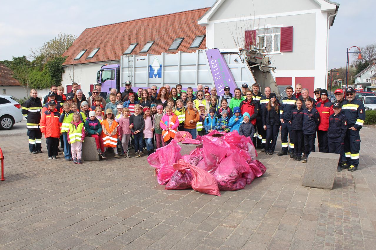A large group of people are posing for a photo in front of a garbage truck, with many children wearing reflective vests and holding pink bags.