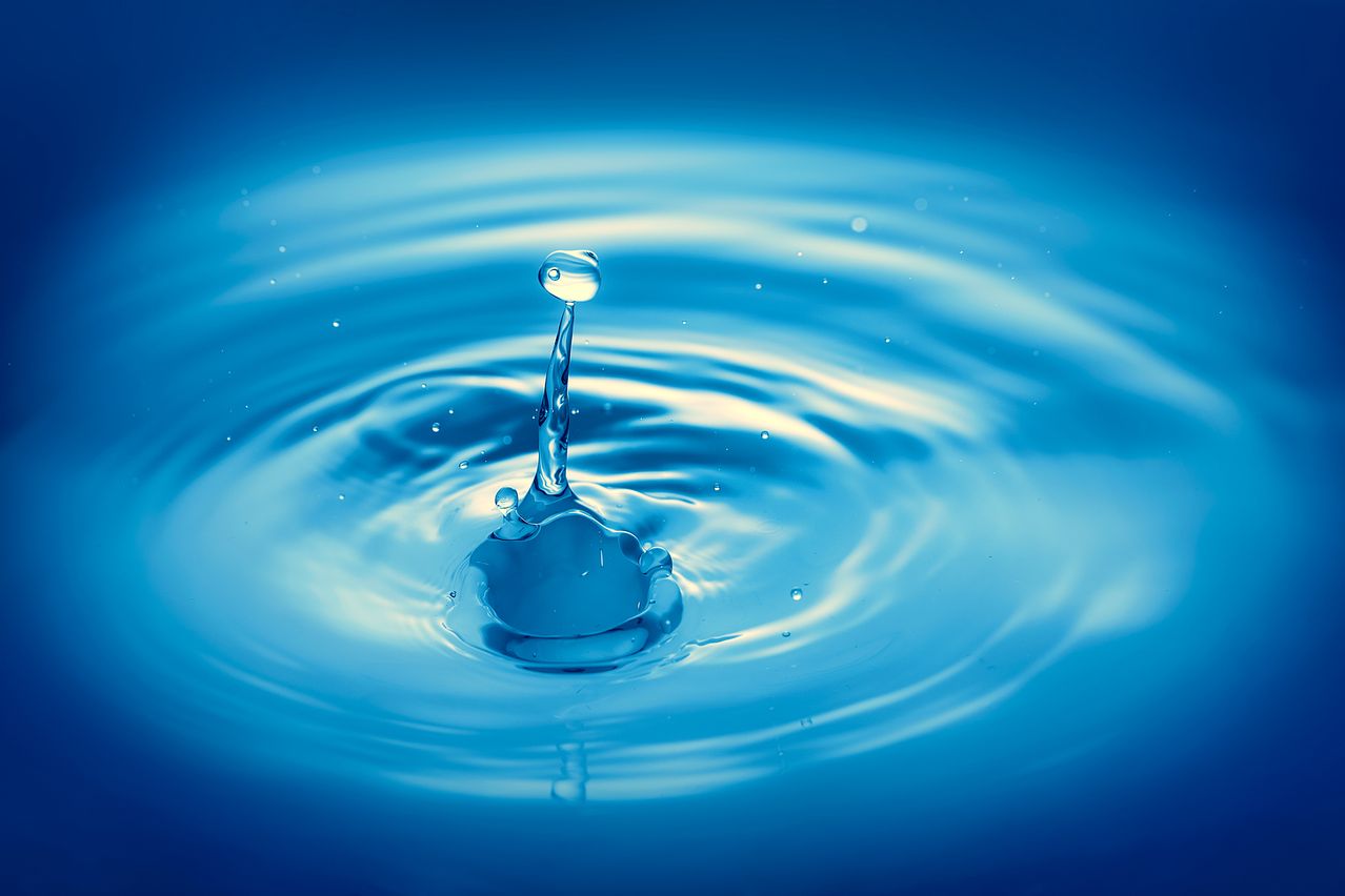 A droplet of water splashes into a pool, creating ripples. The water is clear and blue, reflecting light.