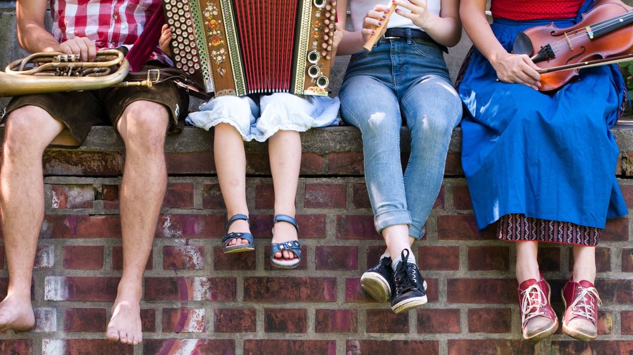 Vier Personen sitzen auf einer Ziegelmauer. Einer spielt Akkordeon, ein anderer Flöte. Ein Mann trägt Shorts und einen Gürtel. Die Frau trägt Jeans und Sandalen.