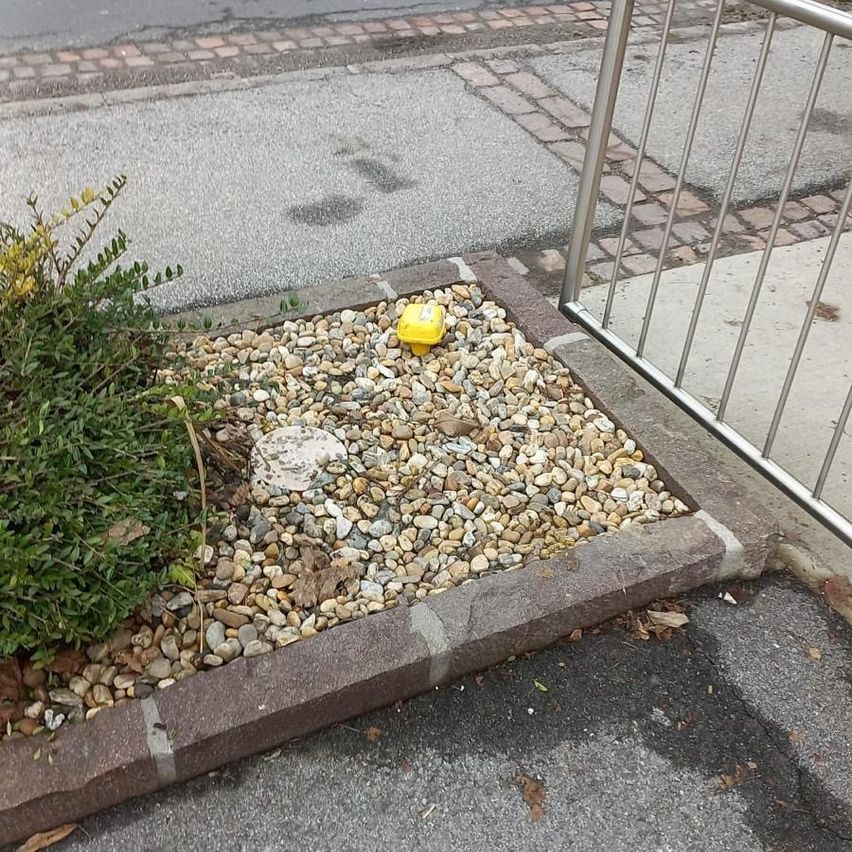 A curb with gravel and a small yellow object sits next to a metal railing on a paved street.