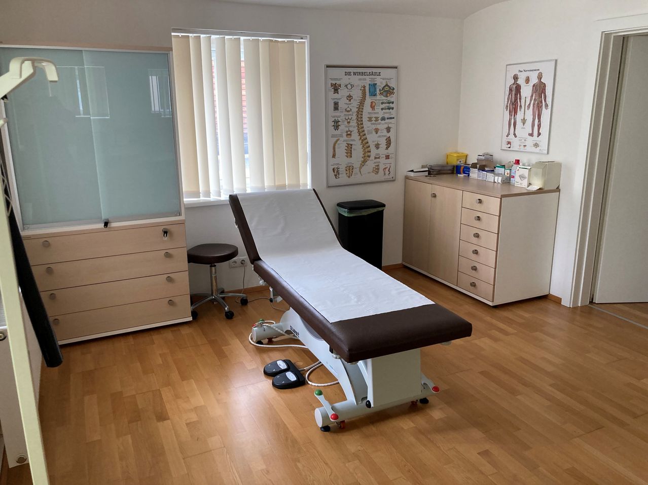 Ein medizinischer Untersuchungsraum mit einem weißen Bett, einem Hocker und einem Schrank mit Schubladen. Ein Poster der Wirbelsäule und eine Glastafel sind an der Wand.
